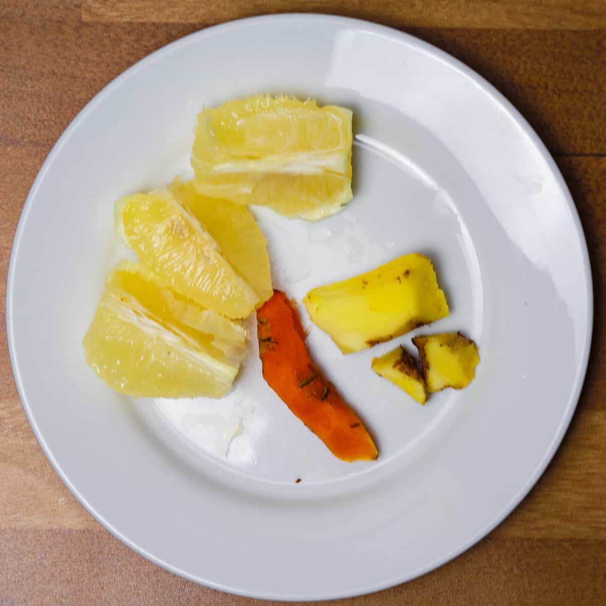 A white plate holding peeled and chopped turmeric, ginger, and sections of lemon and pineapple, prepared for juicing.