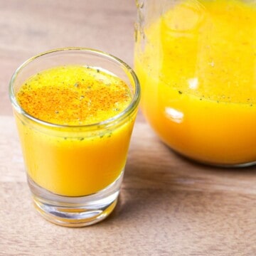 A small shot glass filled with bright yellow anti-inflammatory juice next to a filled mason jar on a wooden table.