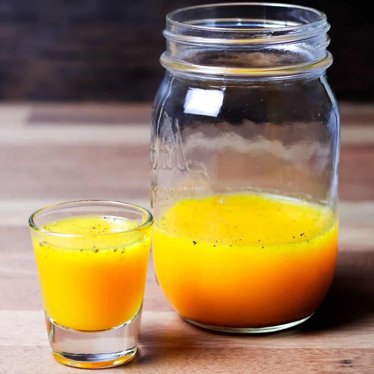 A small glass filled with bright yellow anti-inflammatory juice alongside a larger mason jar half full of the same juice, on a wooden surface.