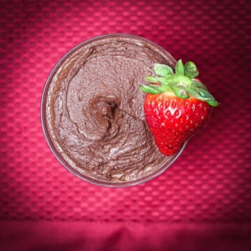 Overhead shot of a bowl of dark chocolate dessert hummus on a red textured background, topped with a whole strawberry, ready to be served.