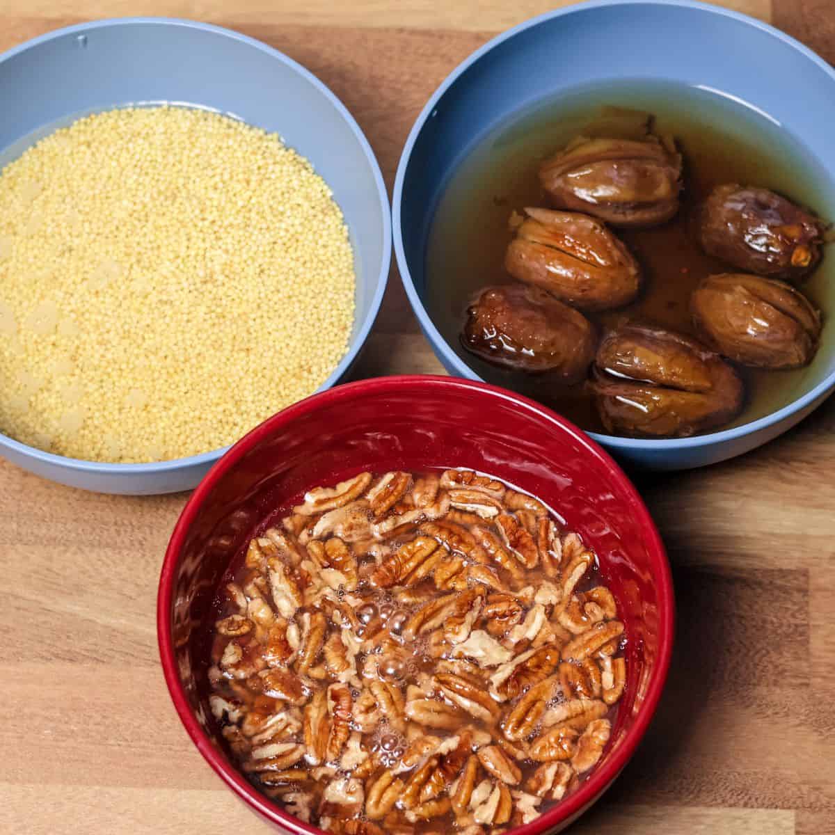 A photograph displaying three bowls: one with yellow millet grains, one with brown Medjool dates in water, and another with pecans in water.
