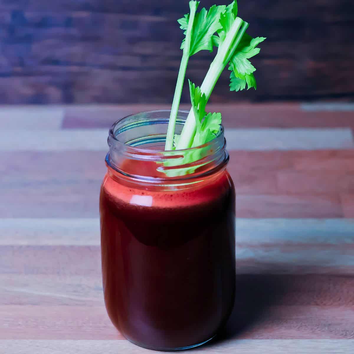 A jar of deep red juice with a stalk of celery as a stirrer on a wooden background, suggesting a freshly made beet-based cleanse juice.
