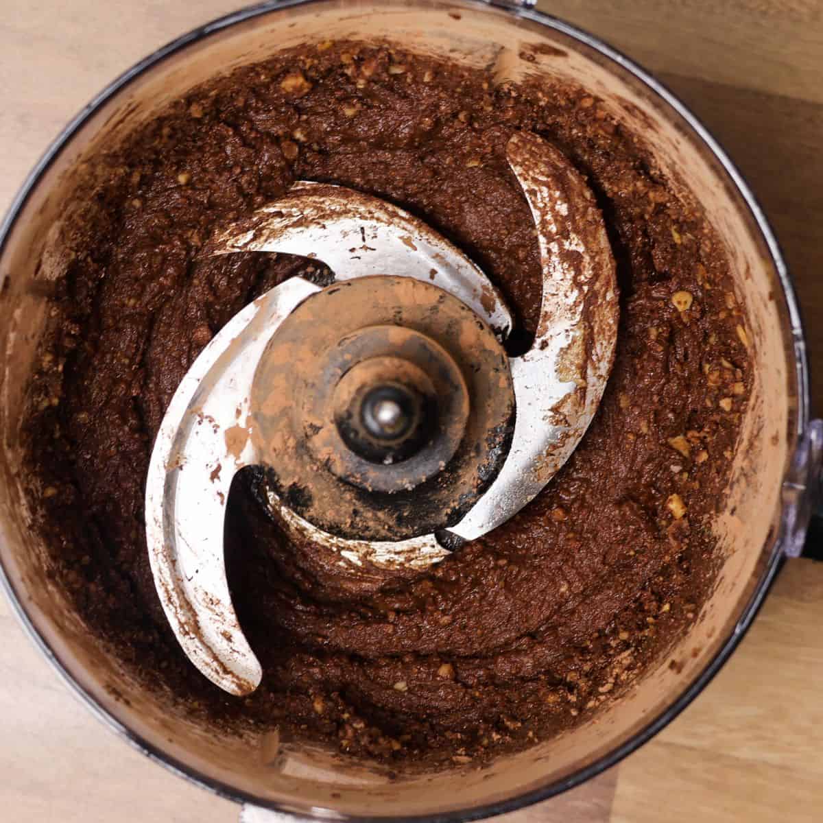 Food processor with dark chocolate hummus ingredients before the addition of almond milk, displaying a thick texture with visible chickpeas.