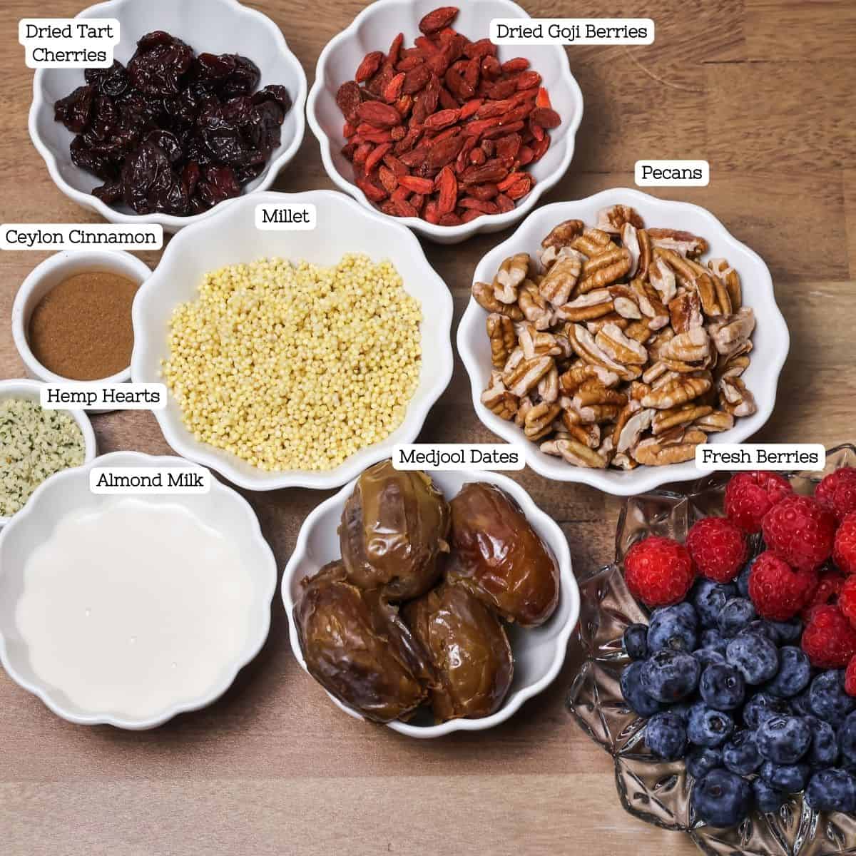 Various ingredients neatly arranged in individual bowls on a wooden surface, including millet, Medjool dates, pecans, goji berries, dried tart cherries, Ceylon cinnamon, hemp hearts, almond milk, and fresh berries.