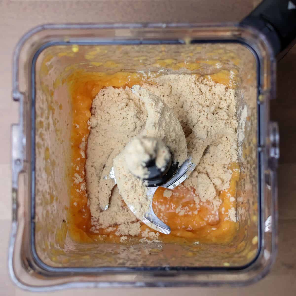 Top-down view of a blender with a layer of vanilla protein powder on top of a base of blended orange fruit mixture.