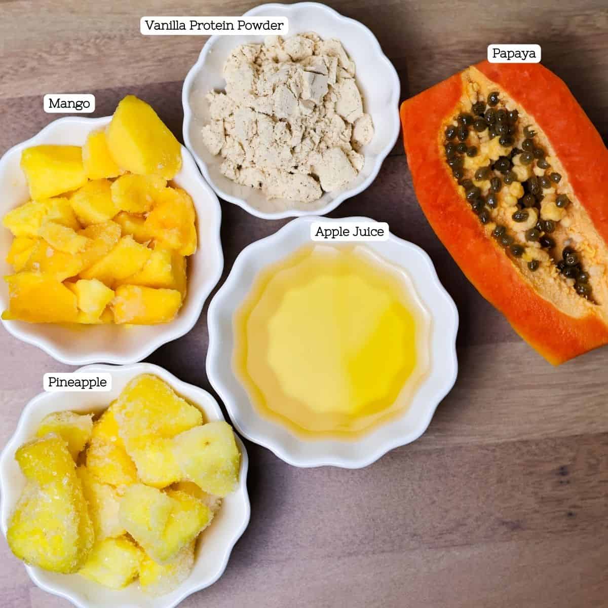 Assortment of ingredients arranged on a wooden surface, including bowls of frozen pineapple, mango, a halved papaya with seeds, apple juice, and vanilla protein powder.