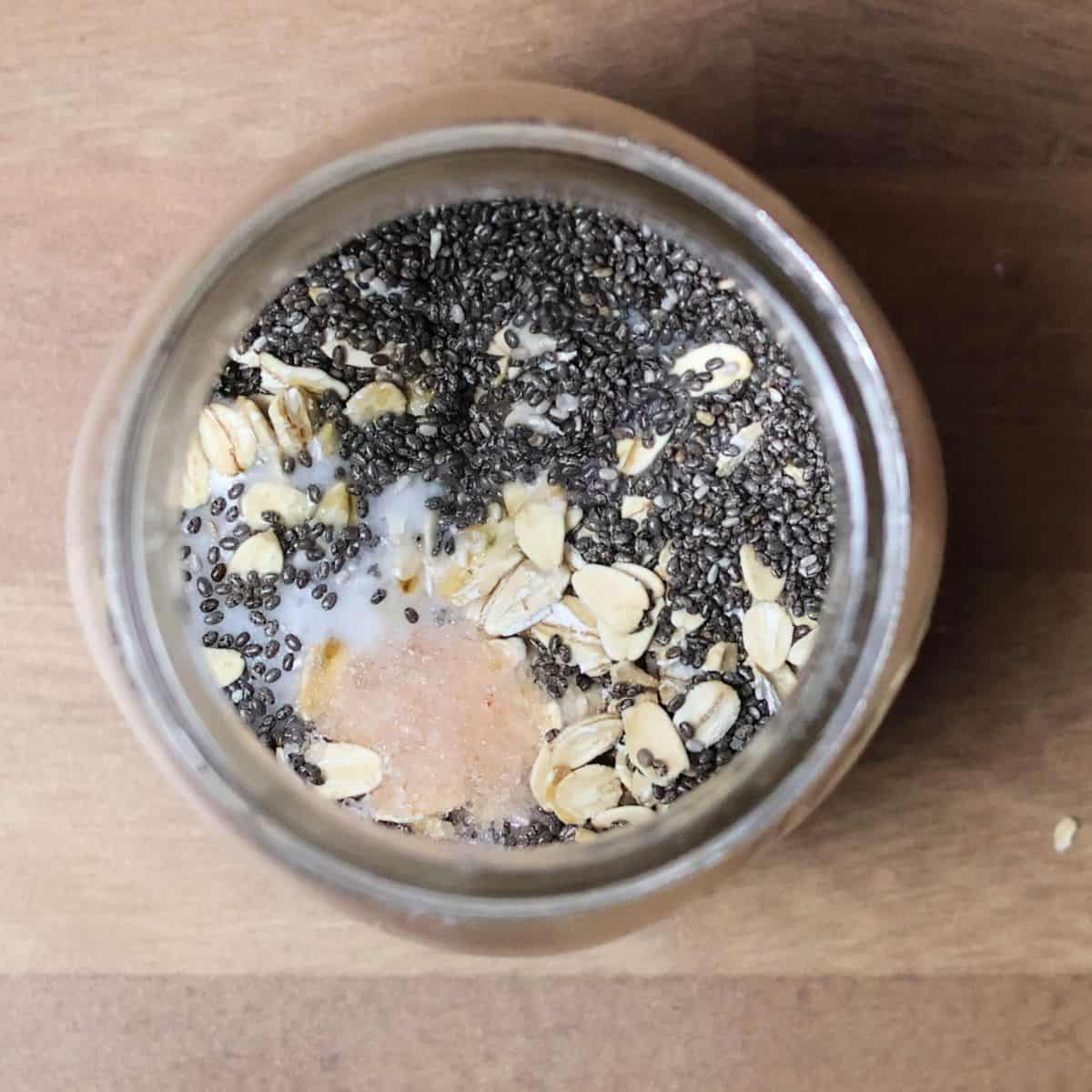 A top-down image of a mason jar showing the initial layer of ingredients for overnight oats before mixing: rolled oats, chia seeds, and a pinch of sea salt, ready to be combined with coconut milk.