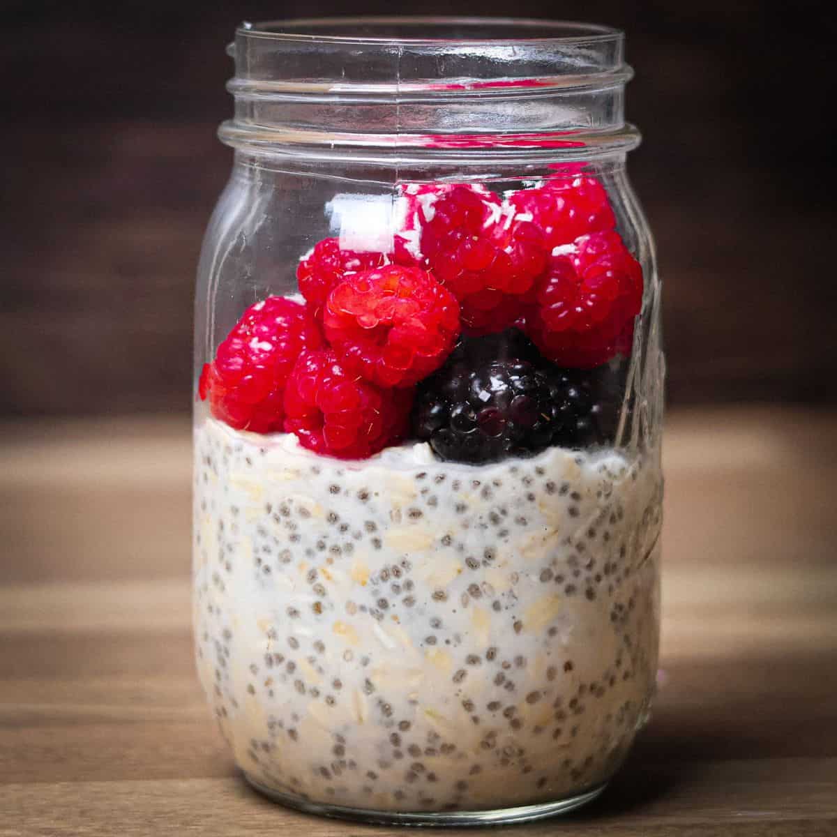 A mason jar filled with overnight oats made with coconut milk and chia seeds, topped with a vibrant heap of fresh red raspberries and a single blackberry, all against a warm, wooden background.