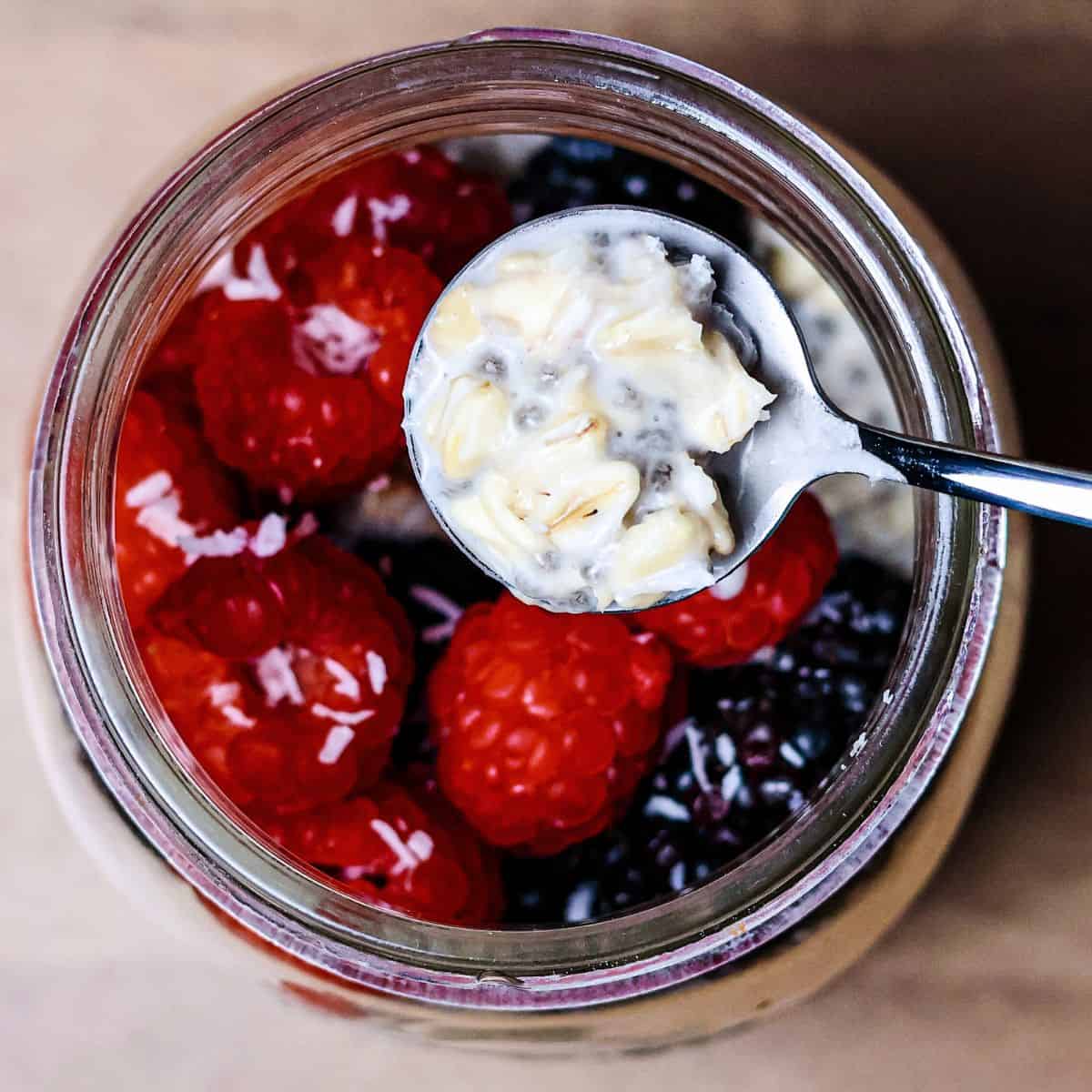 Close-up of a spoon lifting a portion of overnight oats with coconut milk from a jar, revealing the creamy texture and mixed-in chia seeds, with raspberries and blackberries on top.