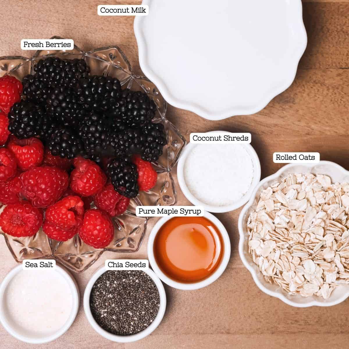 Ingredients for overnight oats arranged on a brown surface, including bowls of fresh blackberries, raspberries, coconut shreds, rolled oats, pure maple syrup, sea salt, and chia seeds next to a jug of coconut milk.