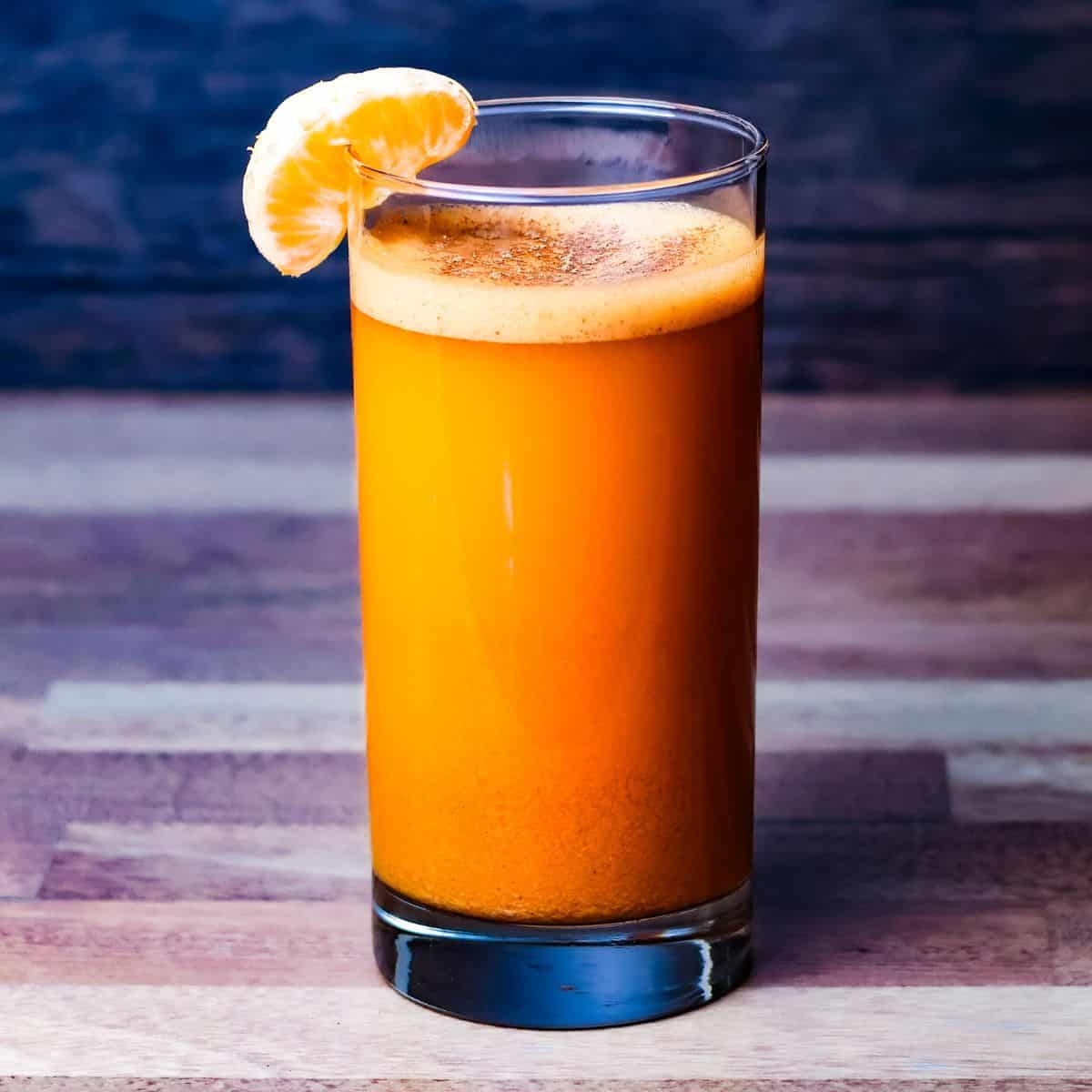 A tall glass filled with rich, orange-colored sweet potato juice, garnished with a bright orange slice on the rim, set against a dark, wooden background. A light frothy layer sits atop the juice, indicating its freshness.