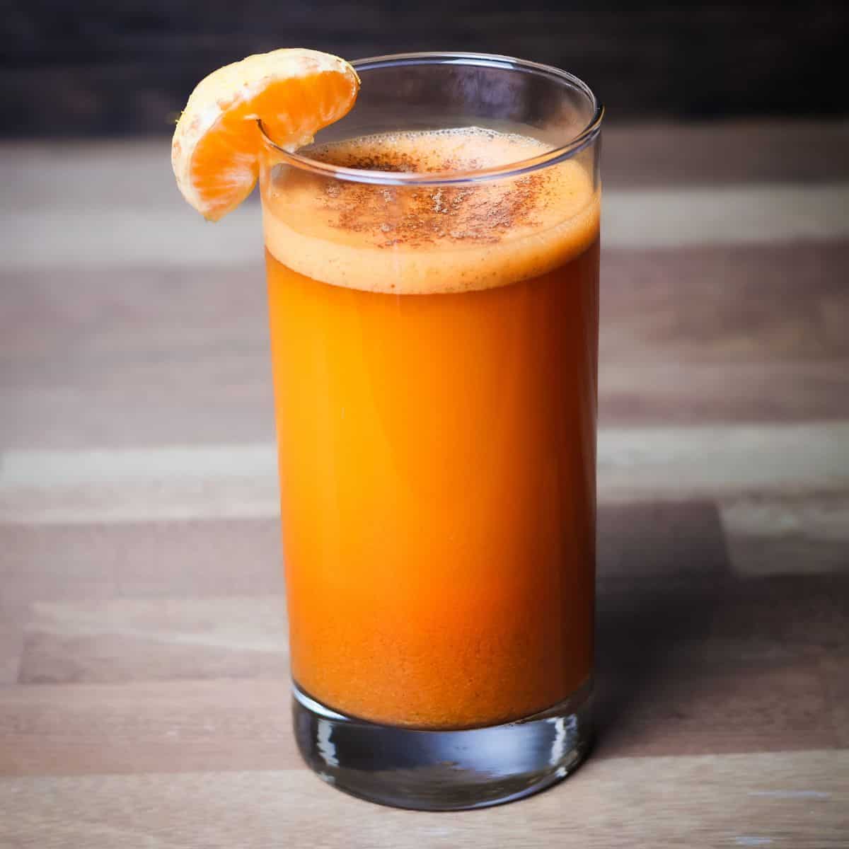 A clear glass filled with freshly made sweet potato juice, exhibiting a deep orange hue. It's garnished with a small slice of orange on the rim, set against a dark wooden backdrop.