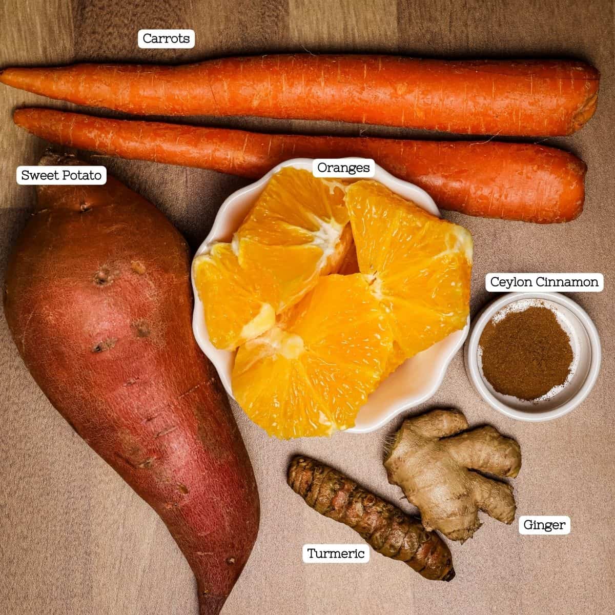 Labeled ingredients for sweet potato juice neatly arranged on a brown surface: whole sweet potato, carrots, orange segments, a piece of turmeric, ginger root, and a bowl of ground Ceylon cinnamon.