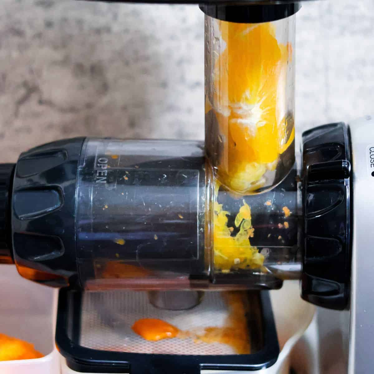 Close-up of a juicer in action, with orange pulp being crushed and juiced, vibrant orange liquid flowing down into the juicer's collection container, against a neutral background.