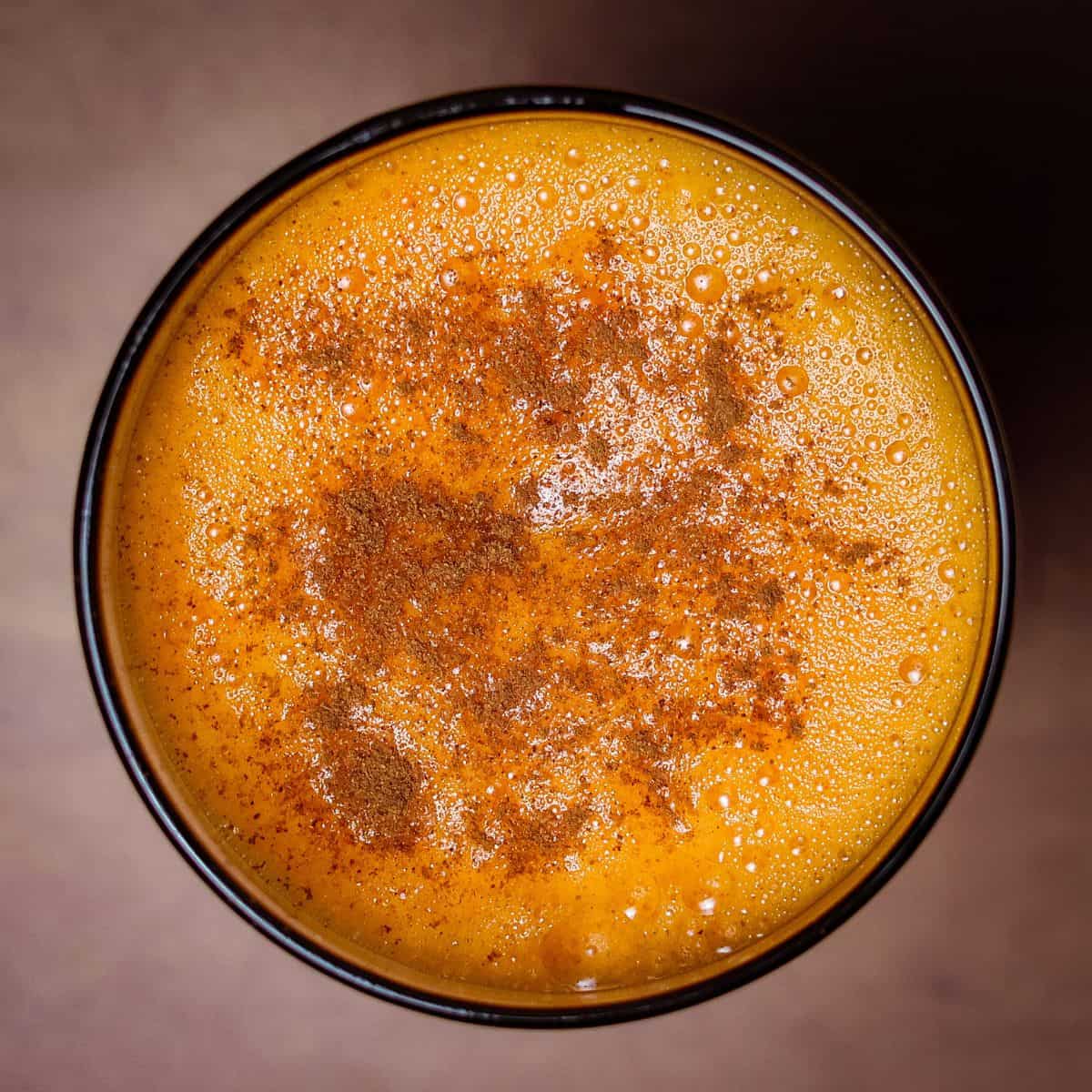 Top-down view of a frothy sweet potato juice in a circular glass, showing a rich texture sprinkled with ground cinnamon, reflecting a golden-orange color.