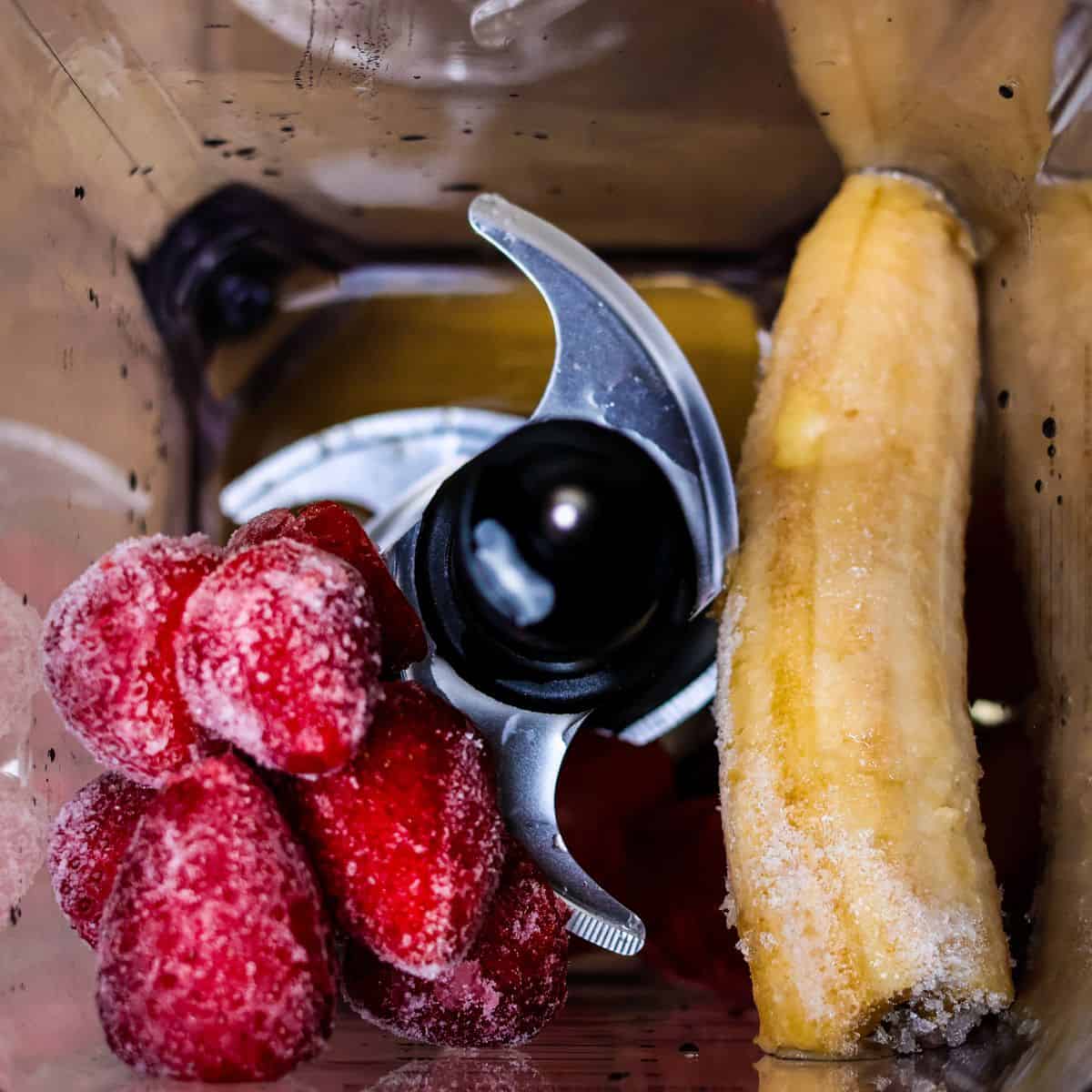 Ingredients ready for blending: frozen strawberries and a banana in the bottom of a blender with a layer of ice crystals on top, indicating they are frozen.