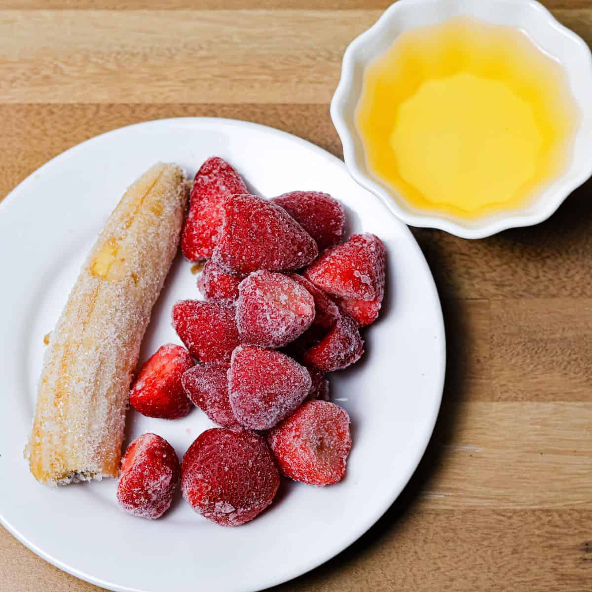 A plate with a whole frozen banana beside a mound of frozen strawberries, with a small bowl of apple juice to the side, ingredients prepped for a smoothie.