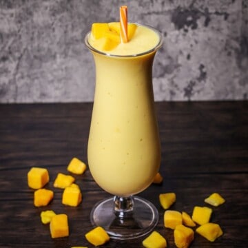 A creamy mango smoothie served in a slender glass, complete with an orange straw and topped with fresh mango pieces, both inside and scattered around the base.