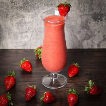 A vibrant strawberry smoothie in a tall glass garnished with a strawberry on the rim, surrounded by fresh strawberries on a wooden table with a gray background.