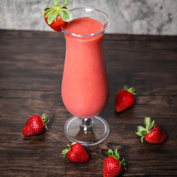 A freshly blended strawberry smoothie in a clear hurricane glass, garnished with a whole strawberry on the rim. The glass sits on a wooden surface with scattered whole strawberries around it, and a muted gray backdrop.