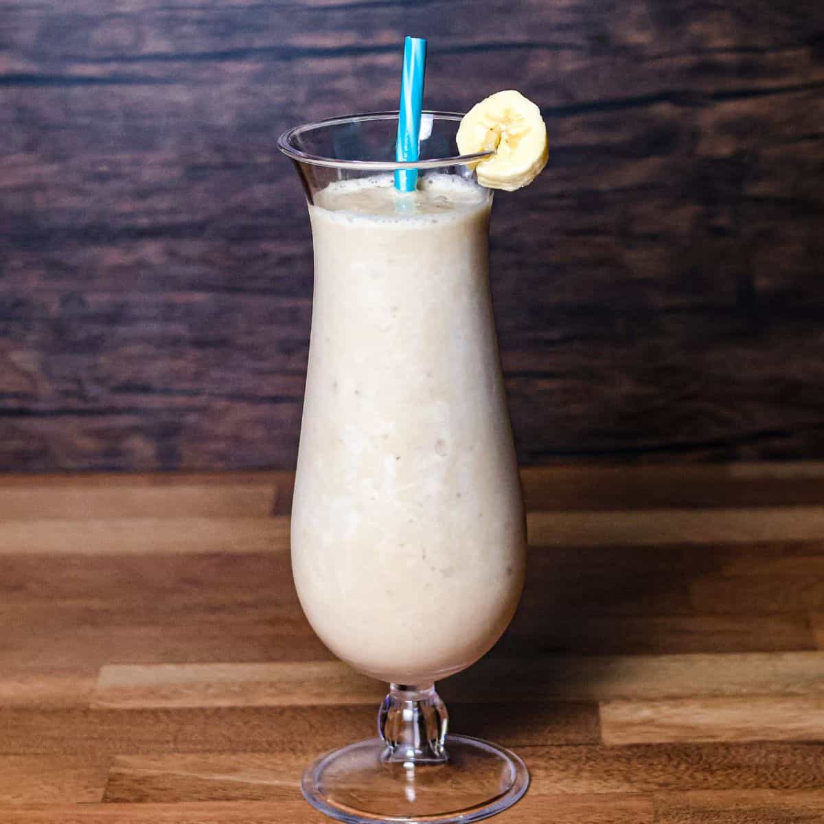A finished banana boat smoothie in a pina colada glass with a blue striped straw and a banana slice garnish on the side.