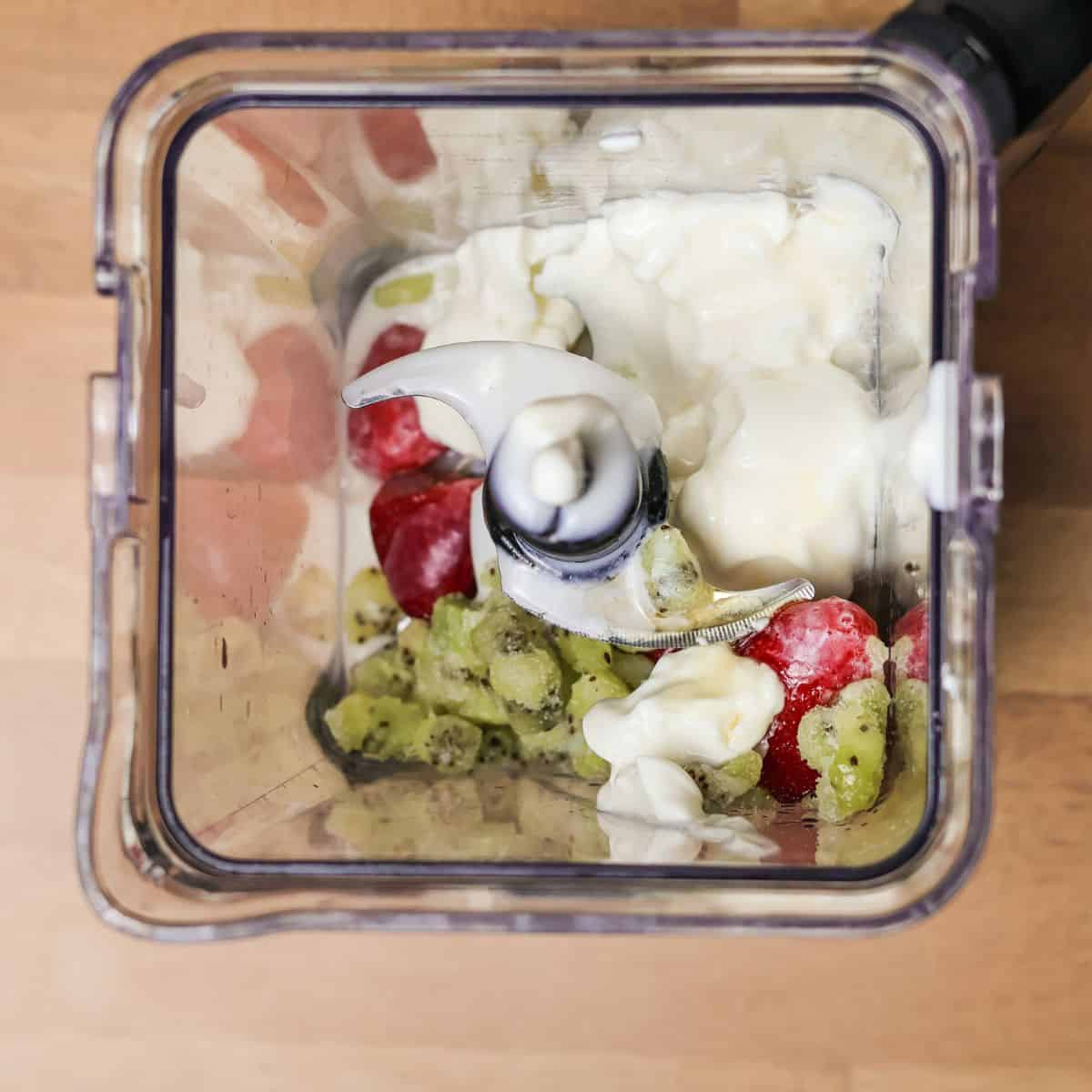 Ingredients for a kiwi quencher smoothie, including frozen strawberries, frozen kiwi pieces, and dollops of Greek yogurt, are placed inside a blender, ready to be mixed.