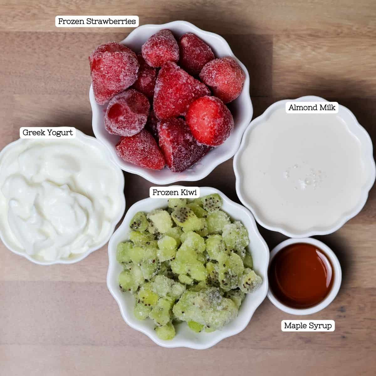Overhead shot of fresh smoothie ingredients neatly arranged on a wooden surface. Clockwise from the top left, there’s a bowl of frosty frozen strawberries, a bowl of smooth almond milk, a small dish of rich maple syrup, and a bowl of vibrant frozen kiwi chunks. In the center, a bowl of creamy Greek yogurt anchors the composition, with labels for each ingredient.