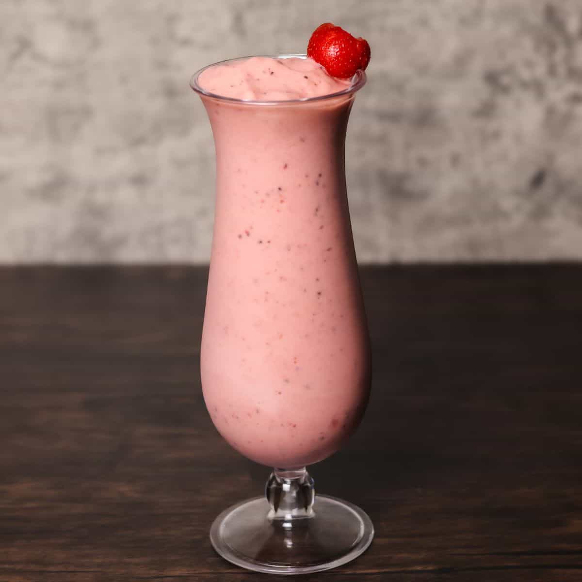 A finished kiwi quencher smoothie served in a tall, frosted glass garnished with a strawberry on the rim, against a dark background.