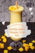 A tall glass of creamy mango smoothie with a vibrant orange hue, topped with fresh mango pieces and an orange straw, with floating white hearts and the text "mindfullyhealthyliving.com Jamba Juice Mango A Go Go" superimposed on a paintbrush stroke.
