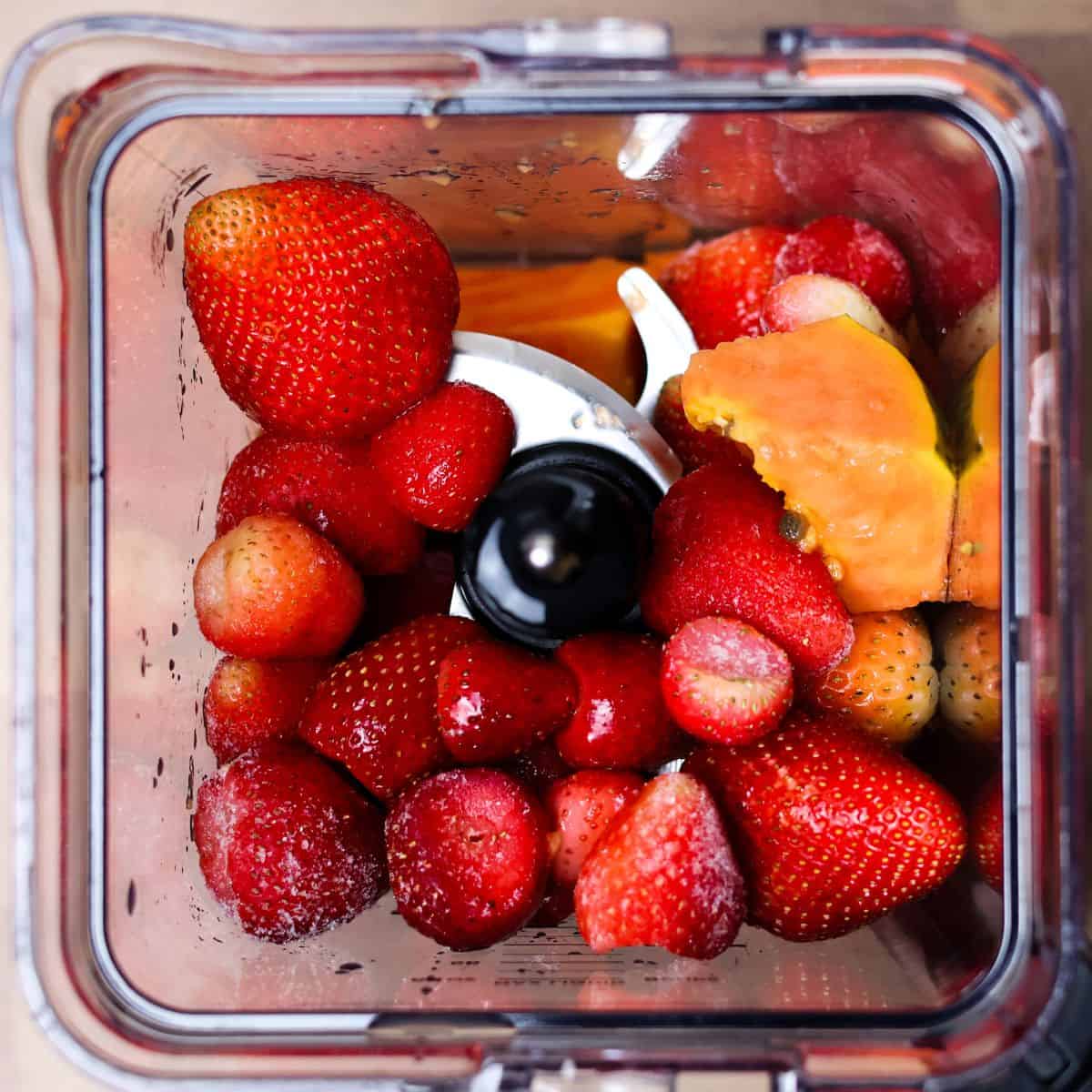 Top-down view of a blender with fresh strawberries, frozen strawberries, chunks of fresh papaya, and a splash of apple juice, ready for blending.