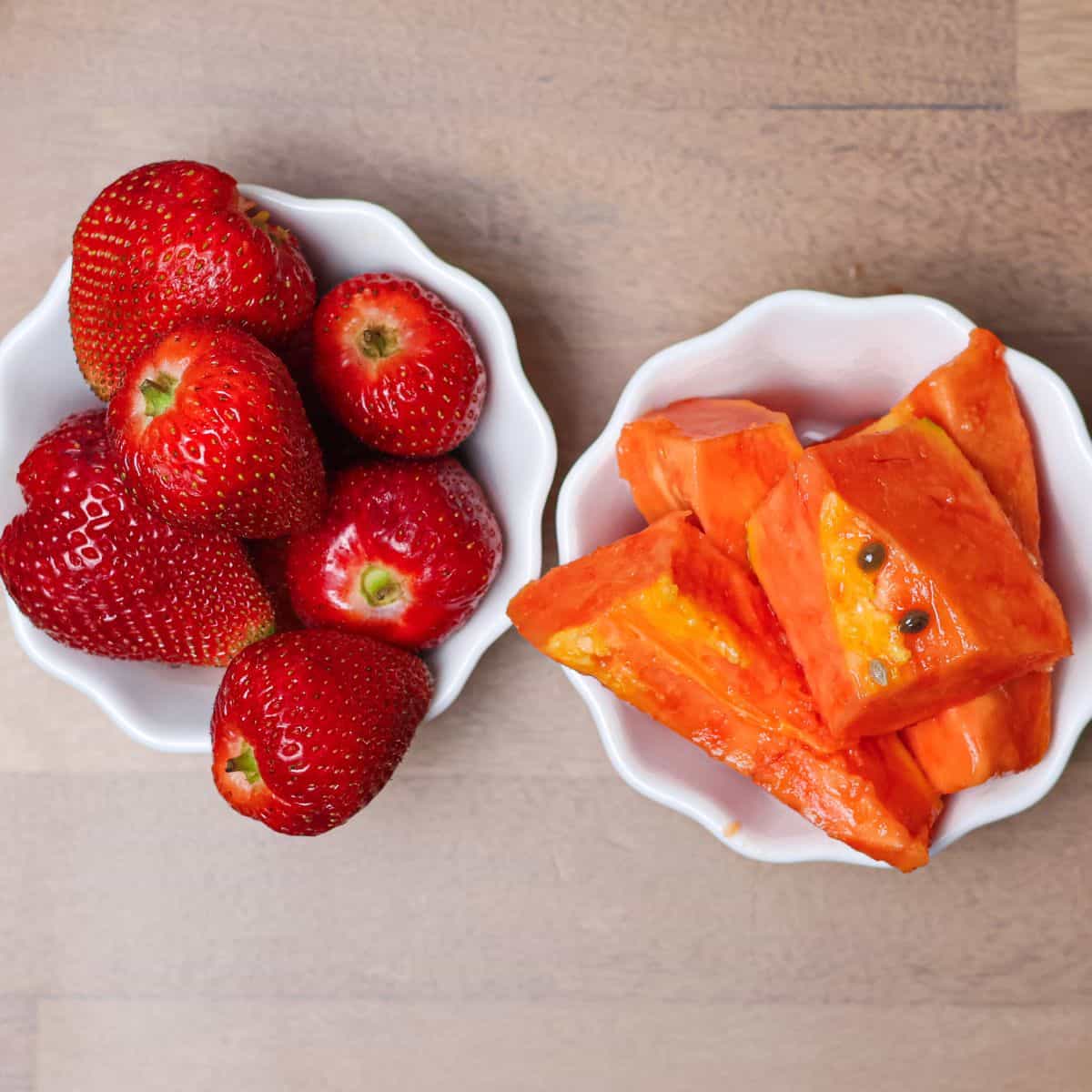 Fresh whole strawberries and freshly cut papaya chunks in separate bowls, prepped for the smoothie.