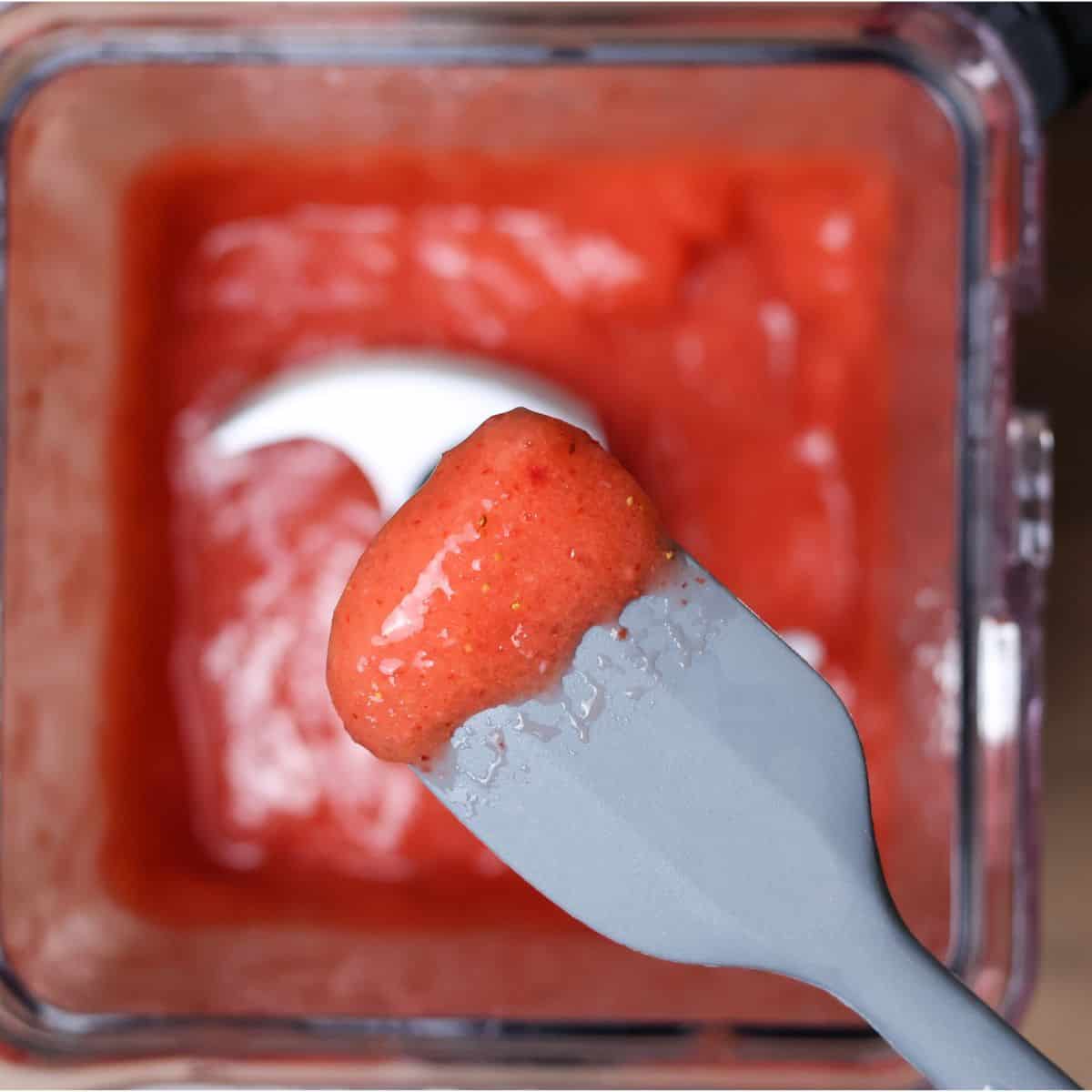 A spatula scooping out a sample of the thick strawberry smoothie from the blender, indicating a taste test for flavor.