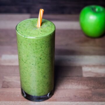 A tall glass filled with a green smoothie, topped with a colorful striped straw, with a green apple in the background, set on a wooden table.