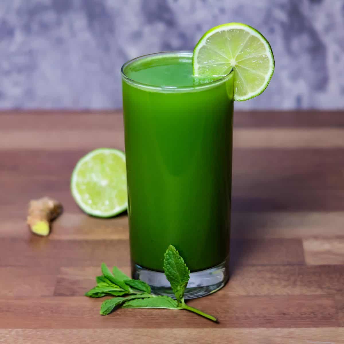 A glass of vibrant green cucumber juice garnished with a slice of lime, with fresh mint leaves, a piece of ginger, and a halved lime in the background.