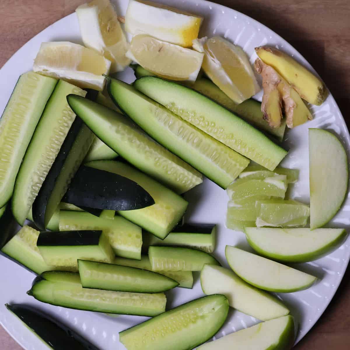 A plate of prepared ingredients including cucumber slices, lemon wedges, lime pieces, ginger slices, and apple slices ready for juicing.