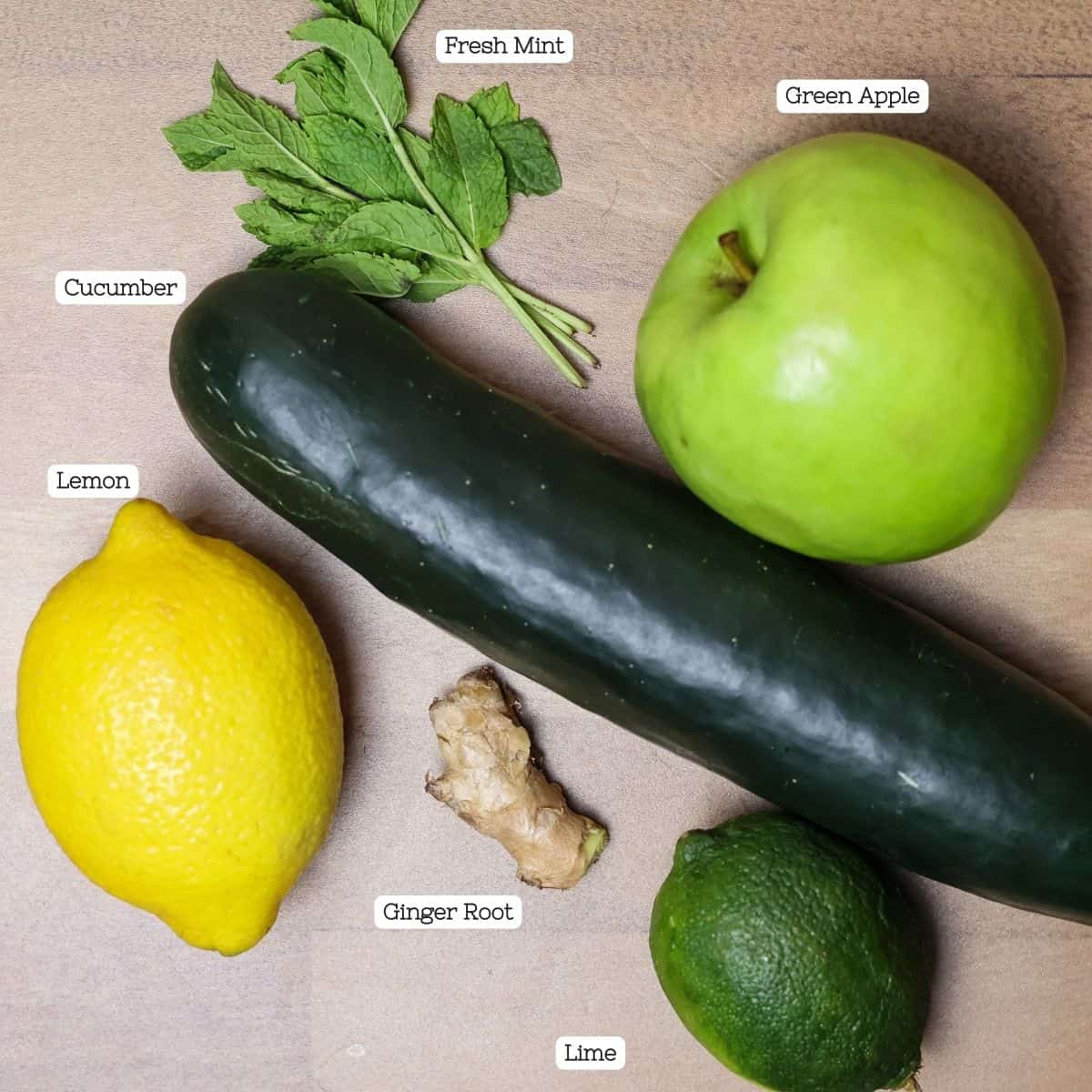 Ingredients for cucumber juice arranged on a wooden surface: a cucumber, a lemon, a lime, a piece of ginger root, a green apple, and fresh mint leaves, each labeled.
