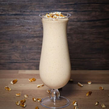 A tall glass filled with a creamy, textured Hulk smoothie garnished with chopped pecans, set on a wooden table surrounded by scattered pecan pieces.
