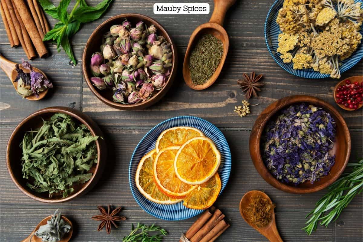 A decorative photo showing various colored rameknins and wooden spoons holding spices and herbs that can go in your tea. Also pictured are slices of orange, dried leaves, anise star, and more.