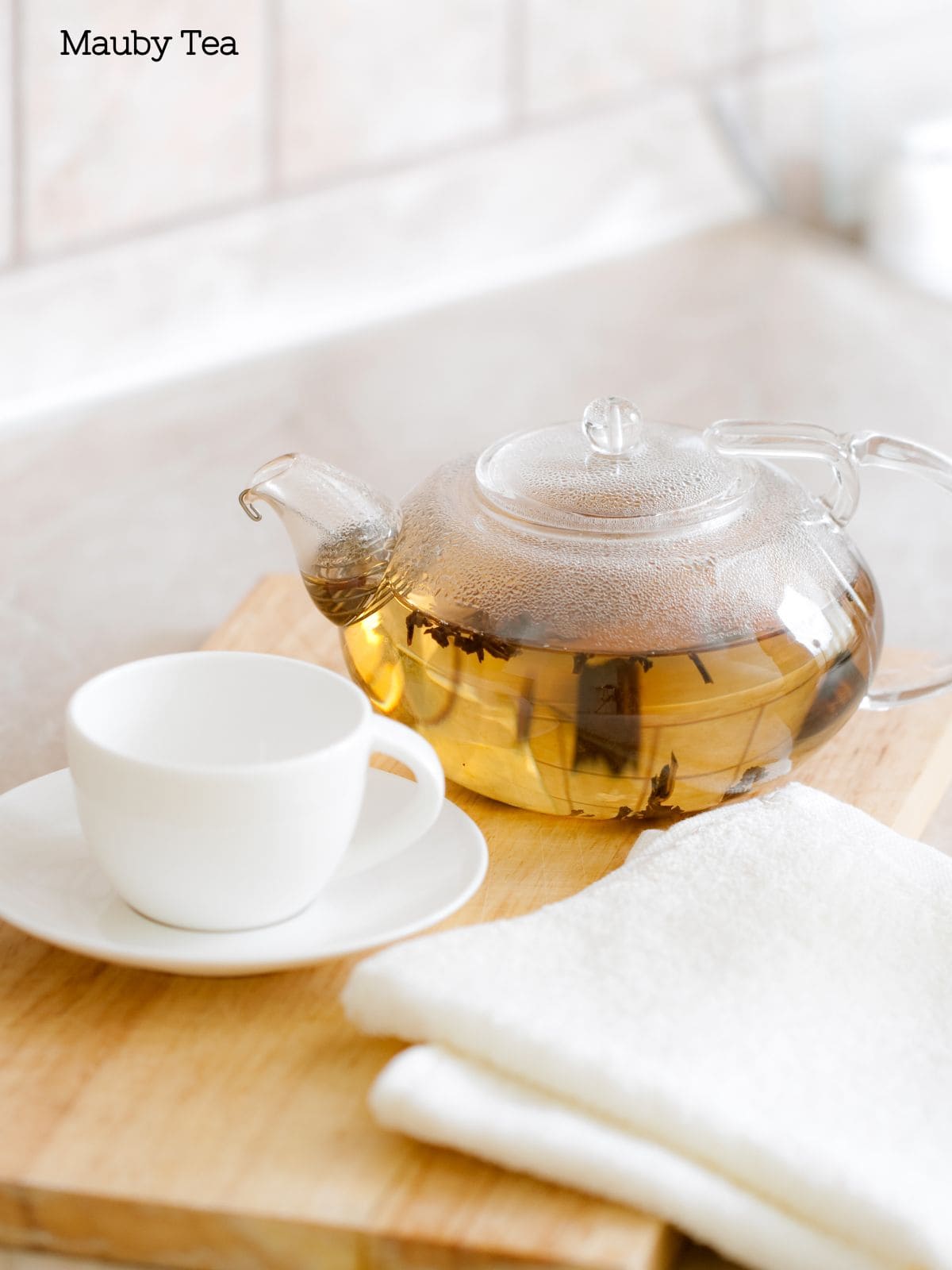 A clear glass tea kettle with light colored Mauby Bark tea in it next to a white teacup on a light wood cutting board and napkin.