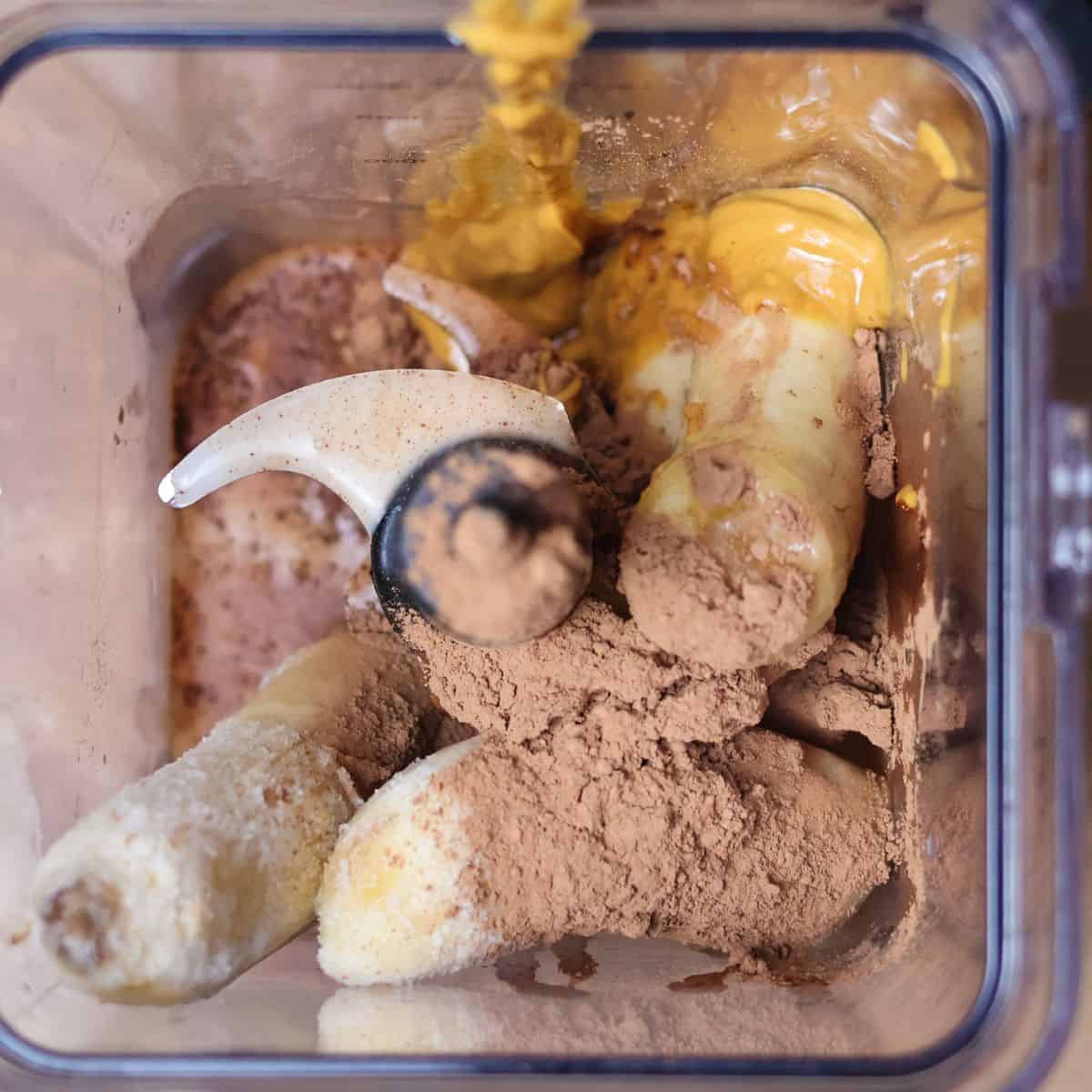 A blender filled with ingredients for a peanut butter banana chocolate smoothie. The ingredients are: sliced bananas, cocoa powder, maple syrup, almond milk, and peanut butter.