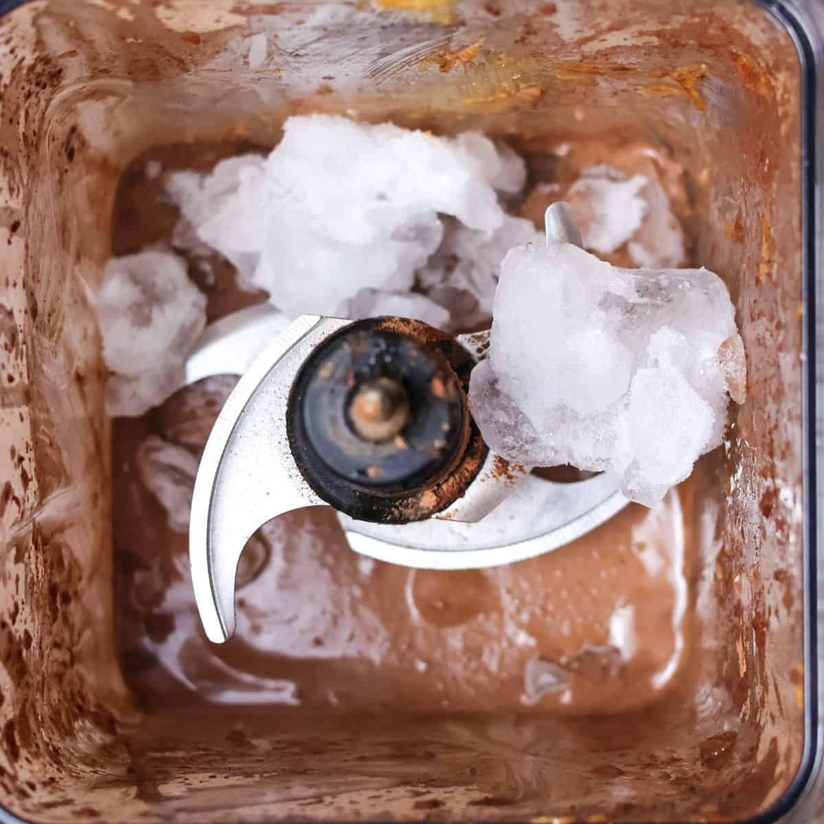 A blender filled with ice cubes on top of chocolate smoothie. The chocolate is swirled throughout the ice, and there is a dark layer of chocolate at the bottom of the jar. It is ready to blend again to thicken the smoothie further.