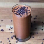 A glass filled with a chocolate smoothie topped with chocolate chips. The smoothie is dark brown and has a thick, creamy consistency. The chocolate chips are semi-sweet and scattered on top of the smoothie.