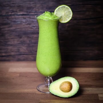 A tall glass filled with a bright green Avocolada smoothie, garnished with a slice of lime on the rim. Next to the glass is a halved avocado on a wooden surface, with a rustic wooden background.