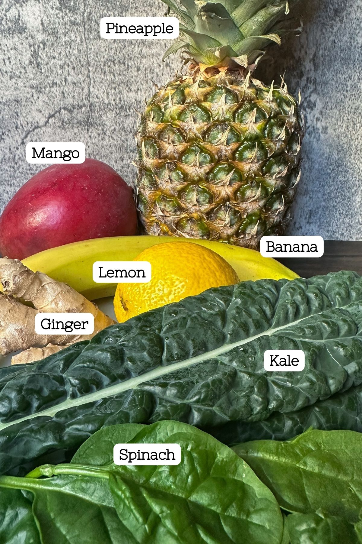 A closeup of the ingredients for detox island green smoothie with labels including spinach, kale, ginger, lemon, banana, pineapple, banana, and mango.