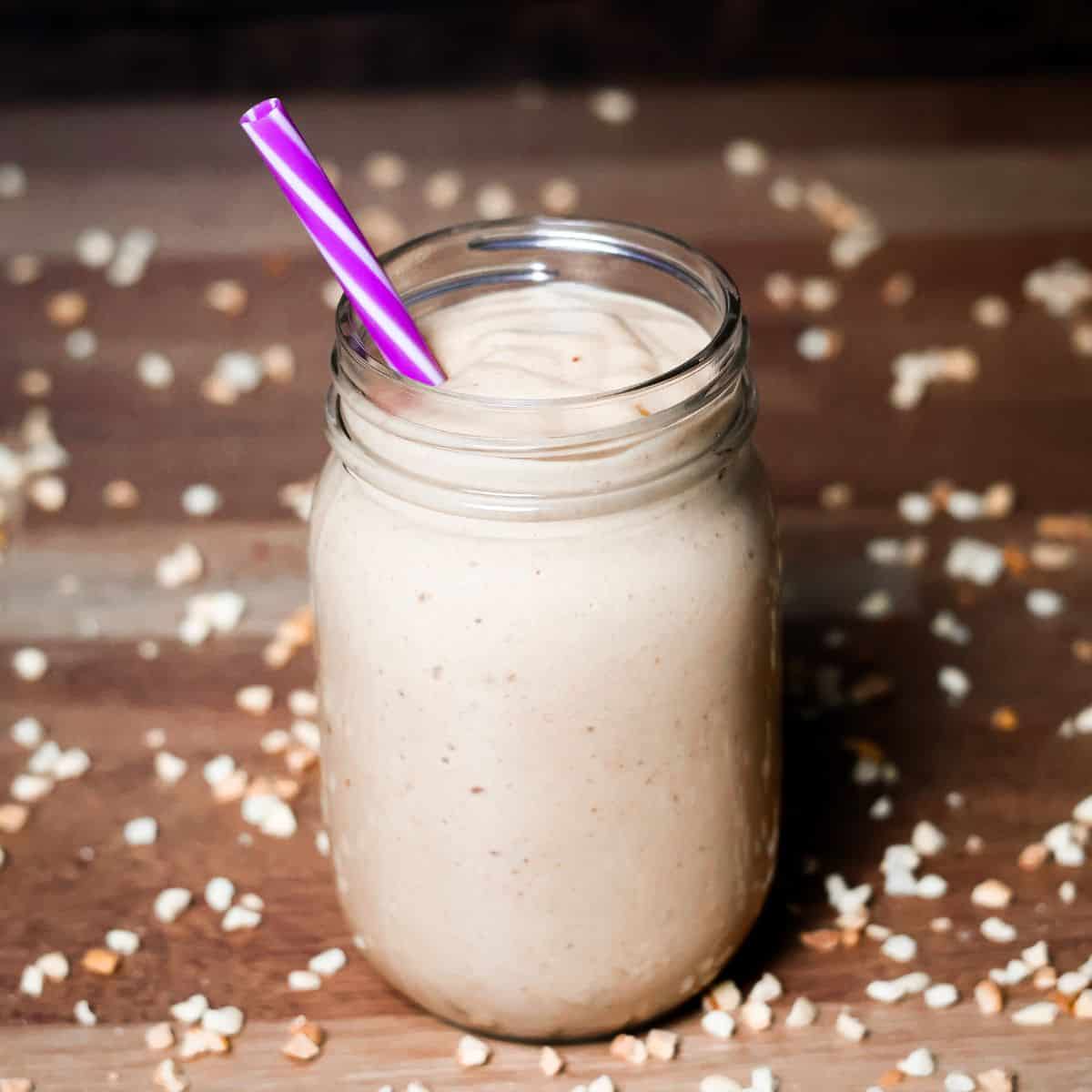 A mason jar filled with a creamy health nut smoothie and a purple-striped straw, surrounded by chopped nuts on a wooden surface.