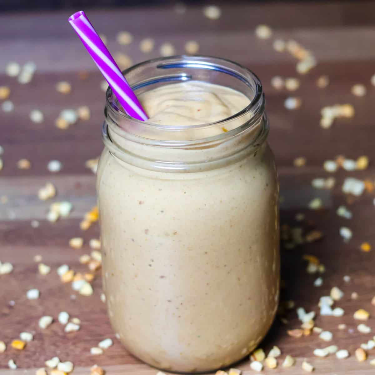 A creamy, light brown smoothie in a mason jar with a purple-striped straw, set on a wooden surface with chopped nuts scattered around.