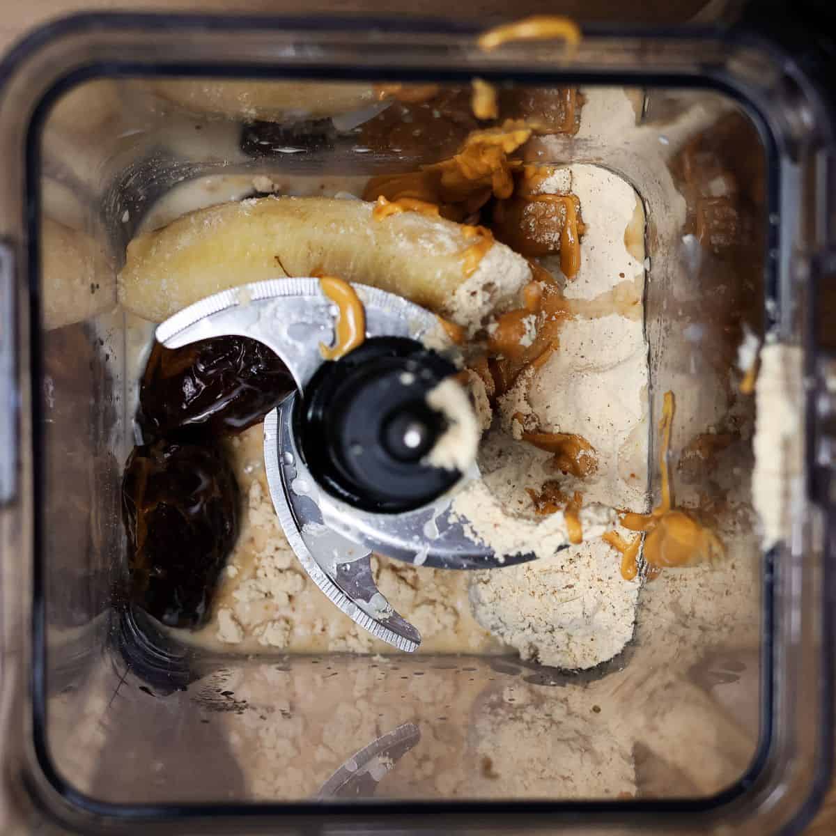 Top view of a blender with ingredients including a frozen banana, pitted dates, peanut butter, and vanilla protein powder, ready to be blended.