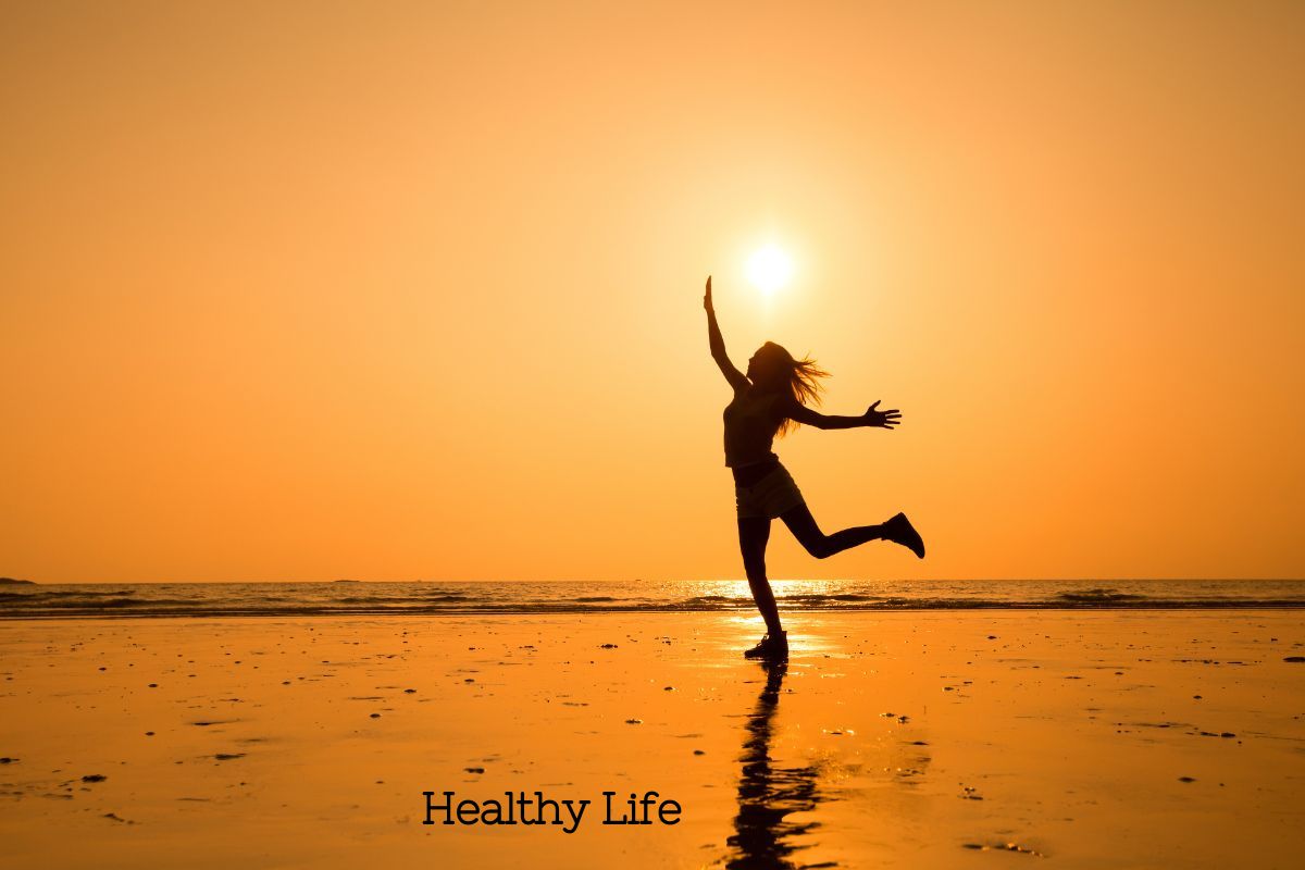 An orange sunset over a beach with a sillouette of a woman dancing under the sun feeling happy and healthy.
