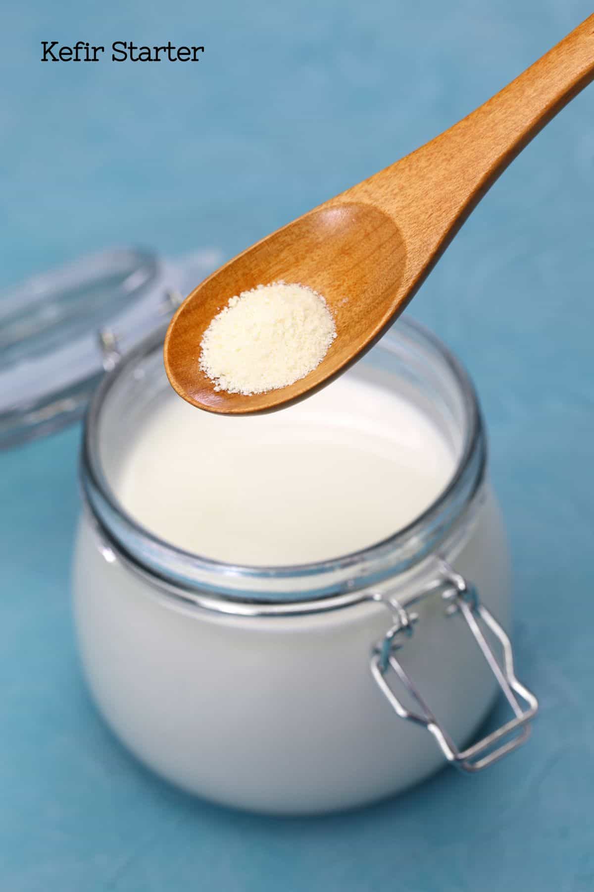 A glass jar of kefir milk with a wooden spoon above it containing powdered kefir starter.