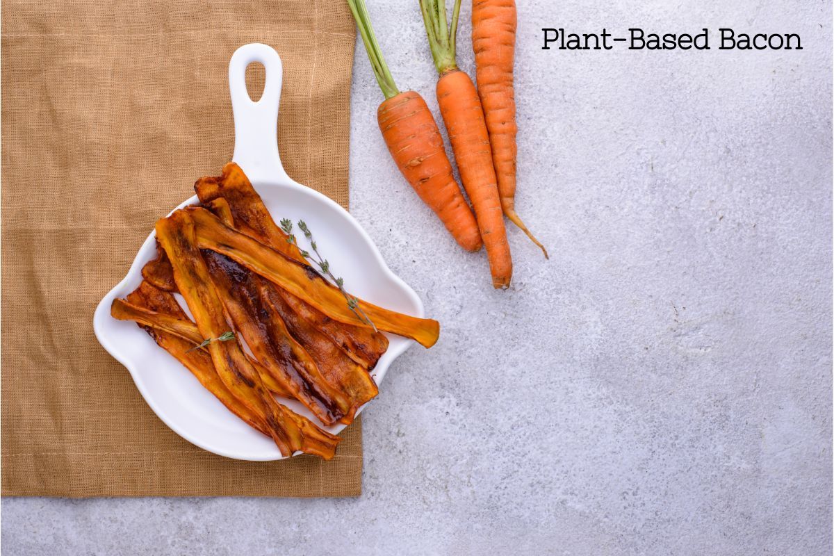 Plant-based bacon made from sliced carrot and cooked with sauces that make them look and taste like the flavors of bacon. The strips are on a white plate next to a group of 3 raw carrots.